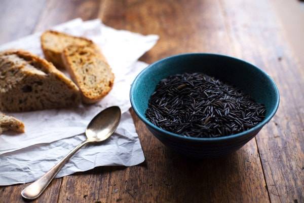 Raw wild rice in a blue bowl with a spoon and bread next to it. 