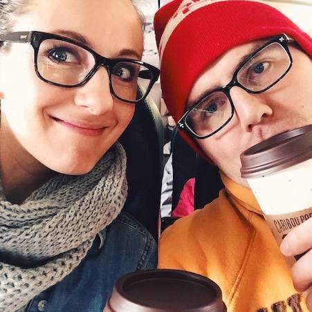 Man in a red cap and a smiling woman in a gray scarf, both holding cups, wearing glasses, and dressed warmly.