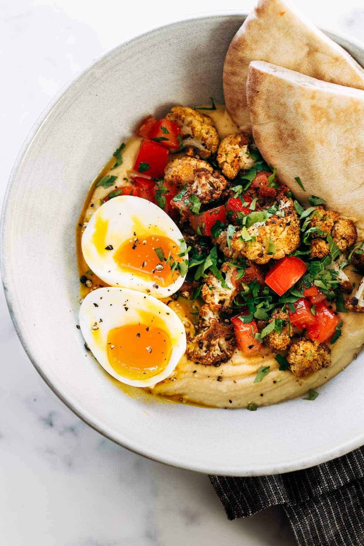 Hummus, roasted cauliflower, eggs and bread in a bowl.