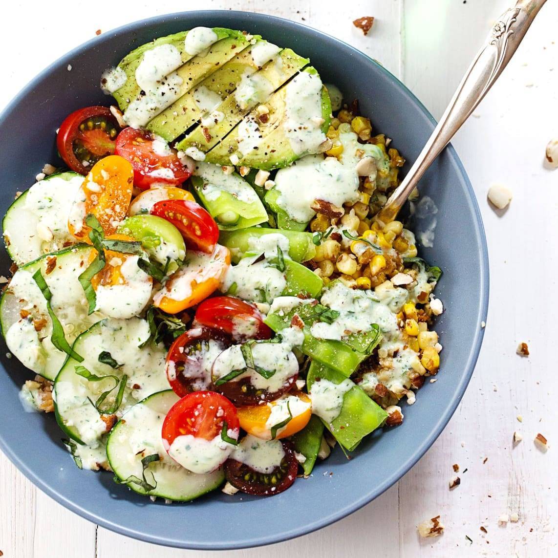 Corn, cucumber, tomatoes, and snow peas, and chopped nuts topped with a dressing in a blue bowl.